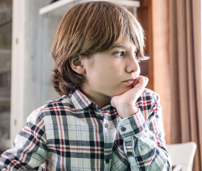 A young boy in a plaid shirt lost in thought indoors during the day.